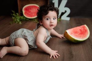 Baby wearing gray romper crawls on wooden floor near large watermelon slices during milestone photo session.