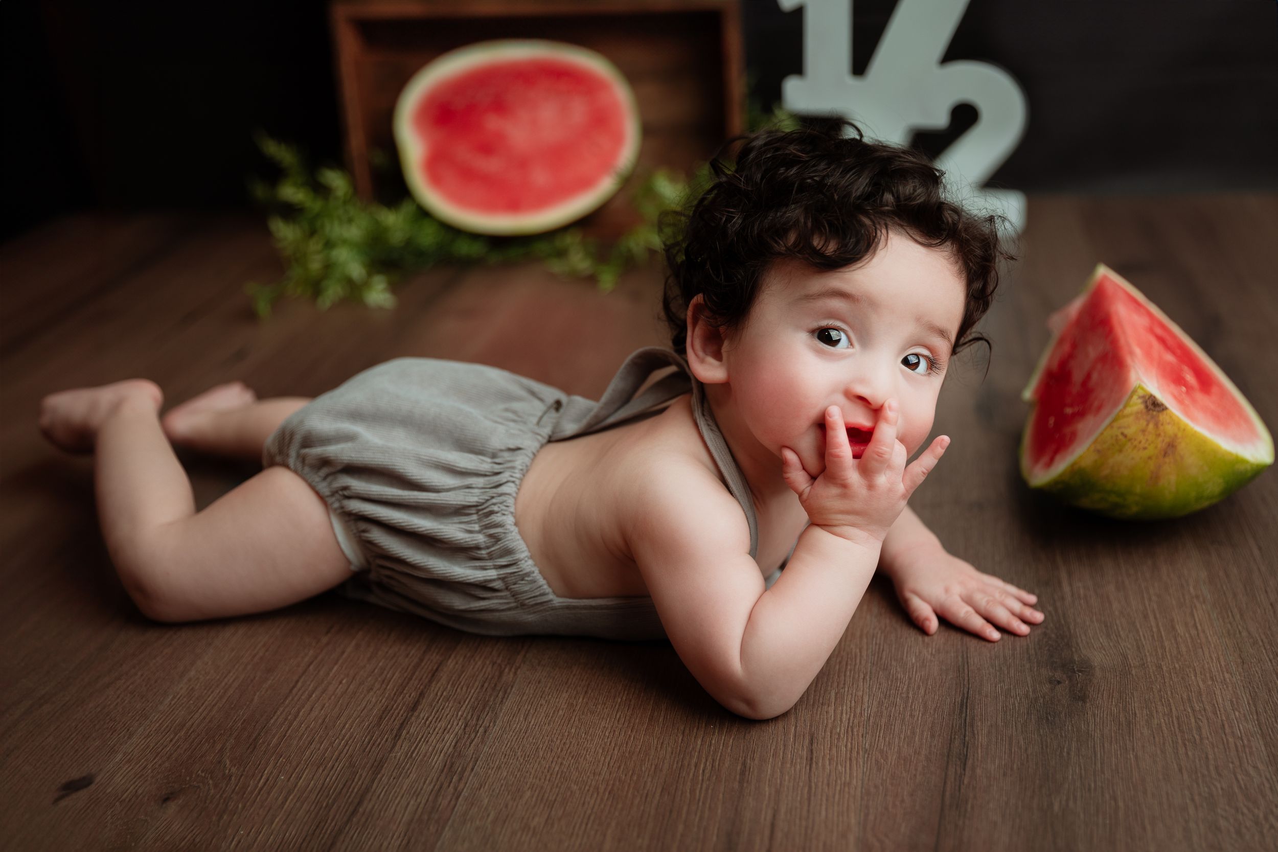 A baby in a gray romper lies on hardwood floor next to sliced watermelon pieces.