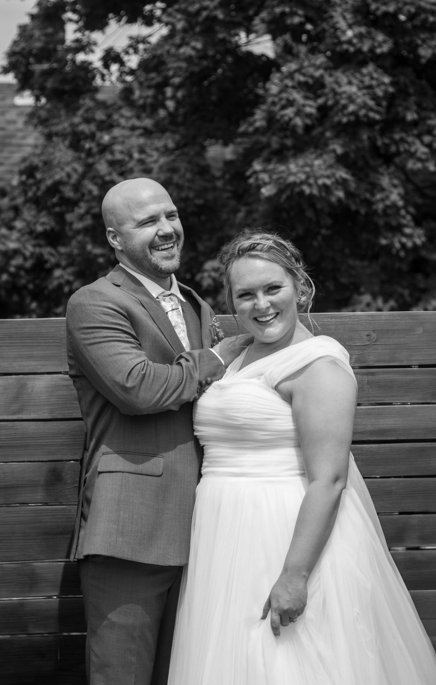 Black and white photo of a happy couple on their wedding day sharing a joyful moment on a wooden bench.