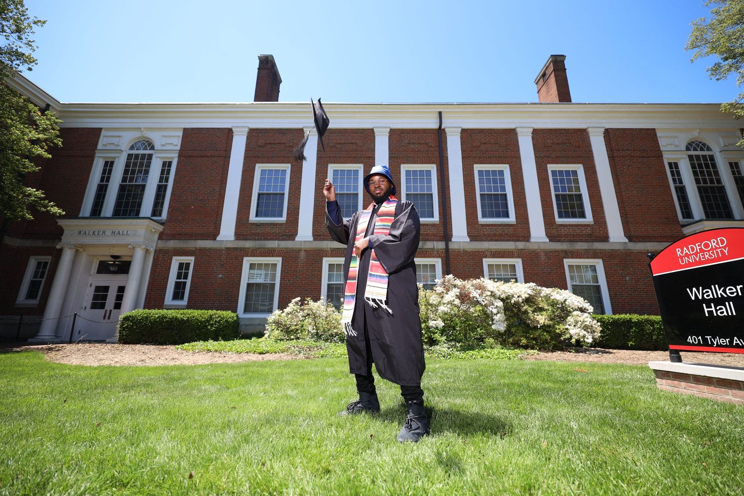 Graduate in regalia posing proudly in front of red brick university building on sunny day.