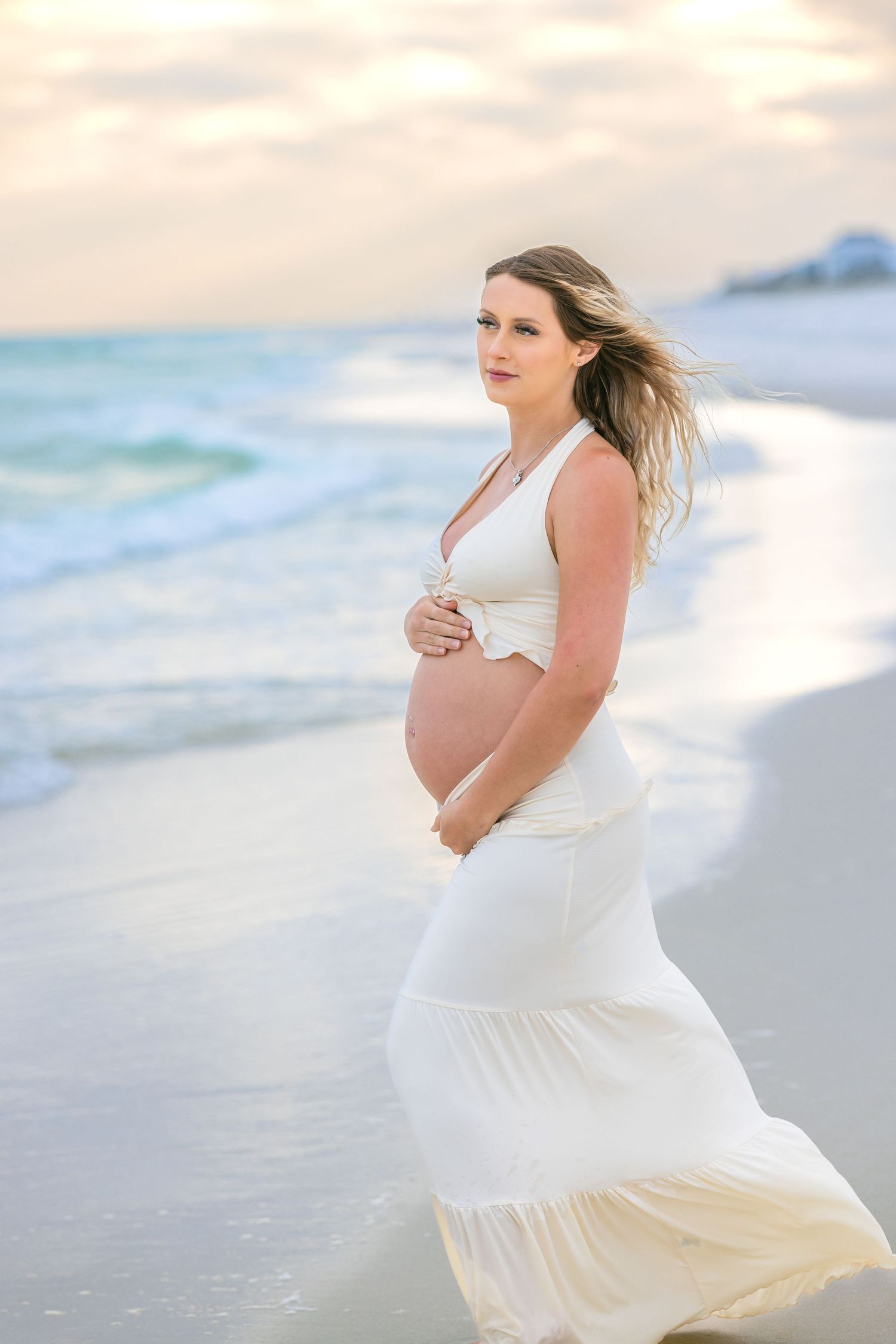 Expectant mother in flowing white dress stands on beach at sunset with waves and cloudy sky in background.