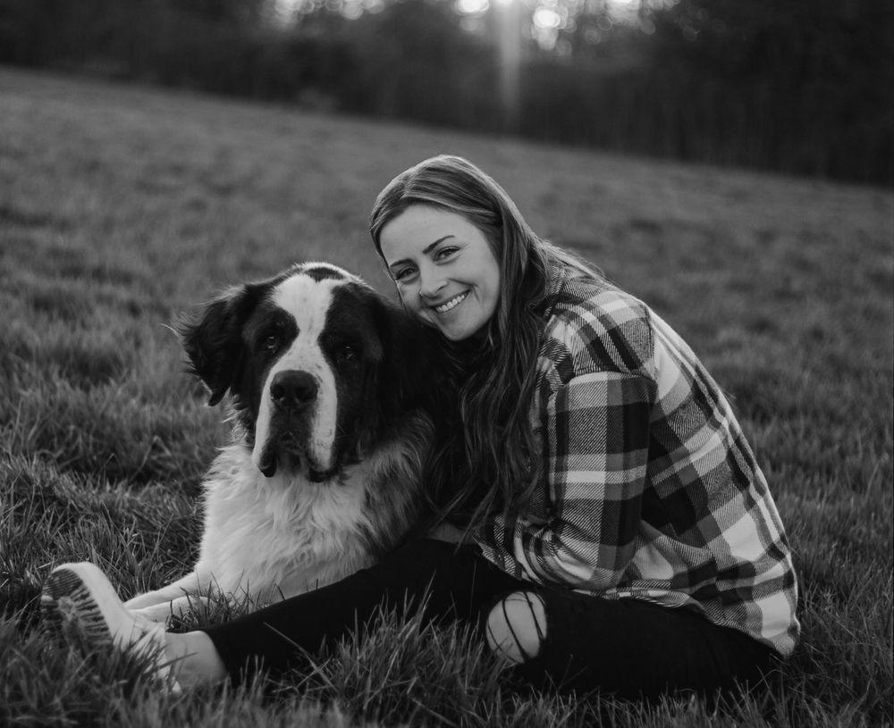 Smiling woman sitting on grass hugging large Saint Bernard dog in black and white photo.