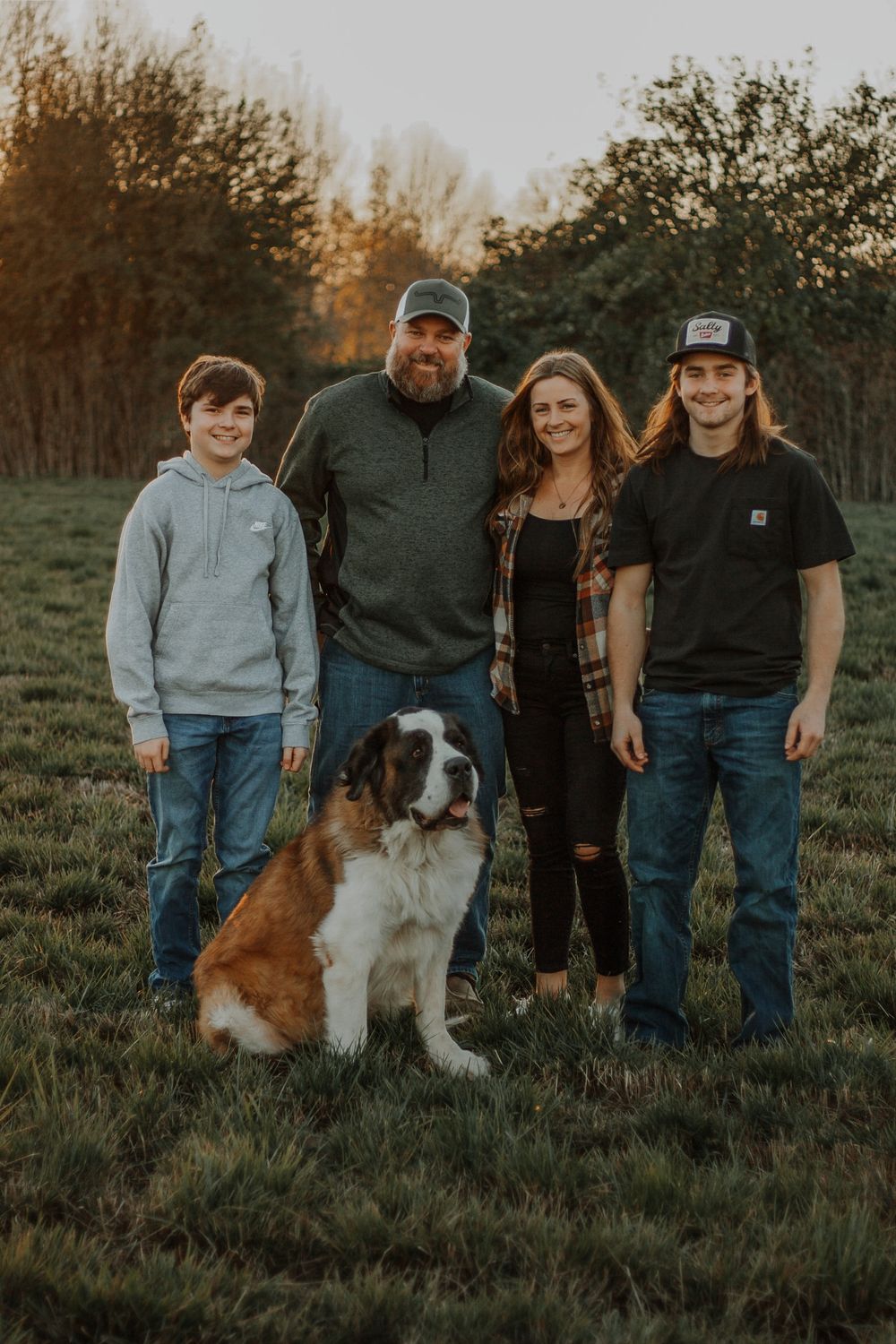 Family of four posing with their Saint Bernard dog in a sunlit green field.