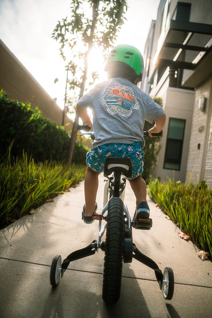 Young child riding a bike with training wheels on a sunny sidewalk, wearing a green helmet and colorful shorts.