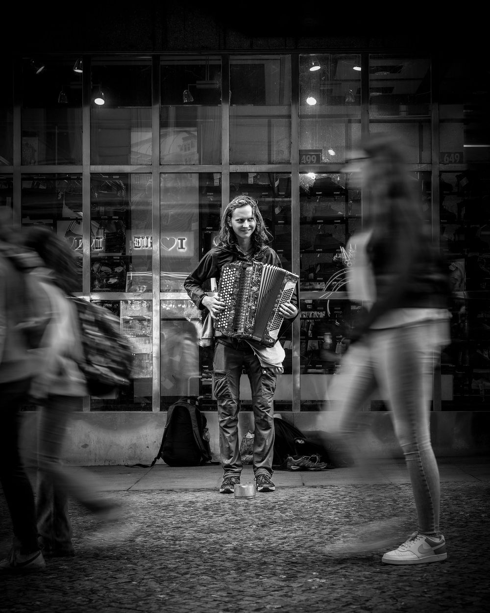 A lone figure stands still on a cobblestone street while blurred pedestrians move past in black and white.