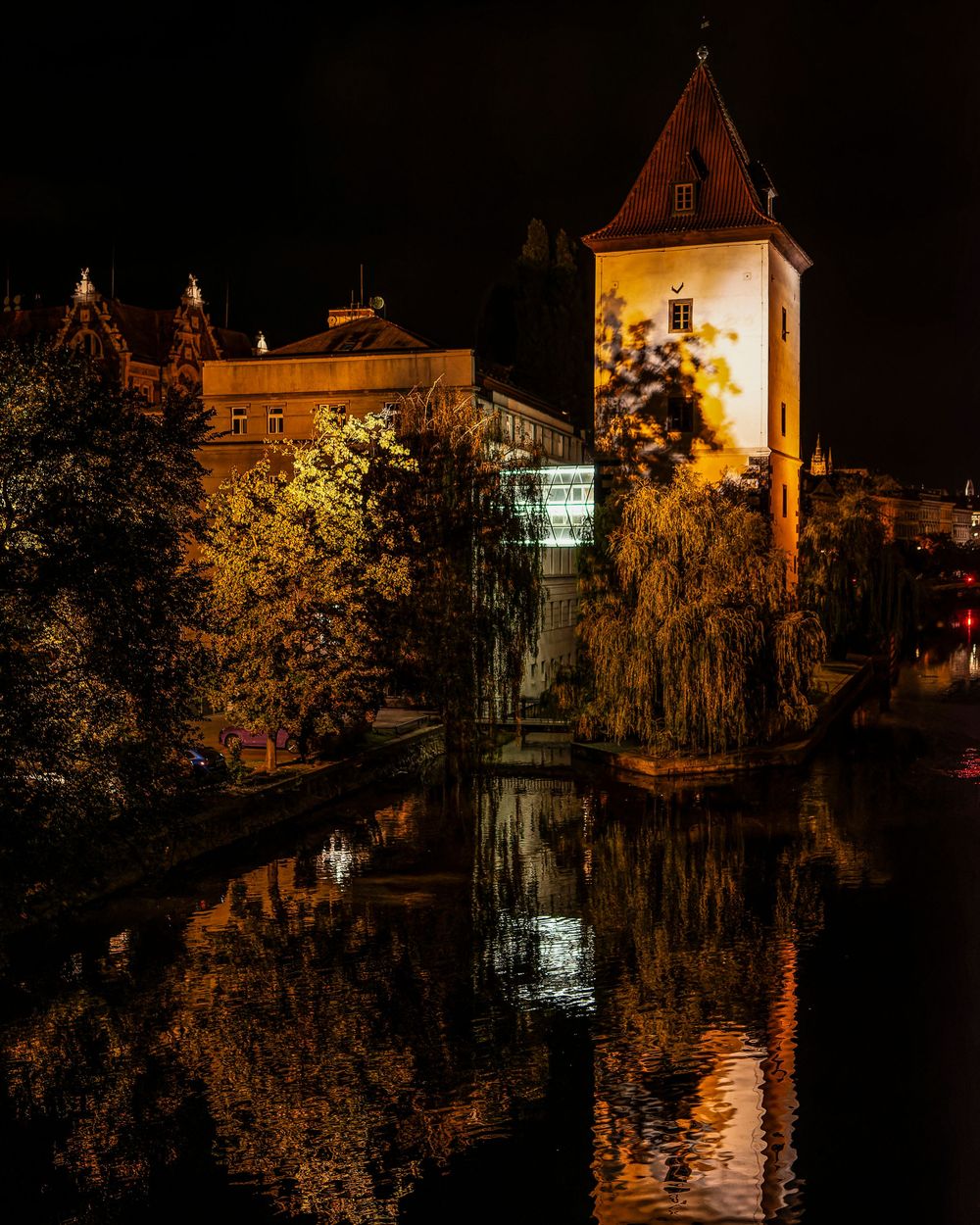 Historic buildings and castle structures illuminated at night reflect beautifully on calm water in a scenic evening view.