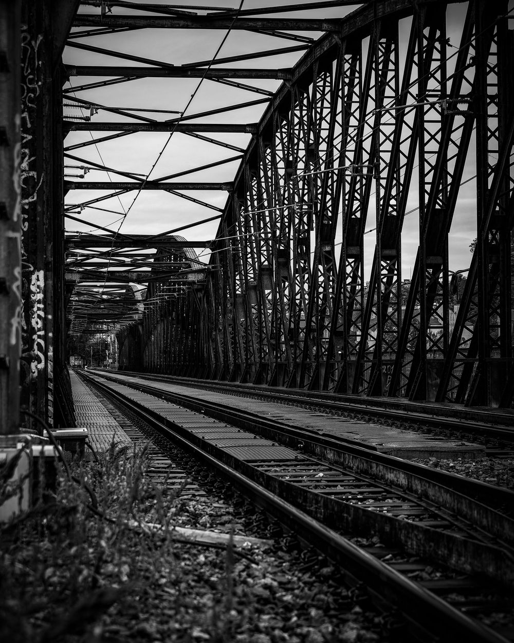 Black and white composition showing railway tracks crossing an industrial steel bridge structure with geometric patterns.