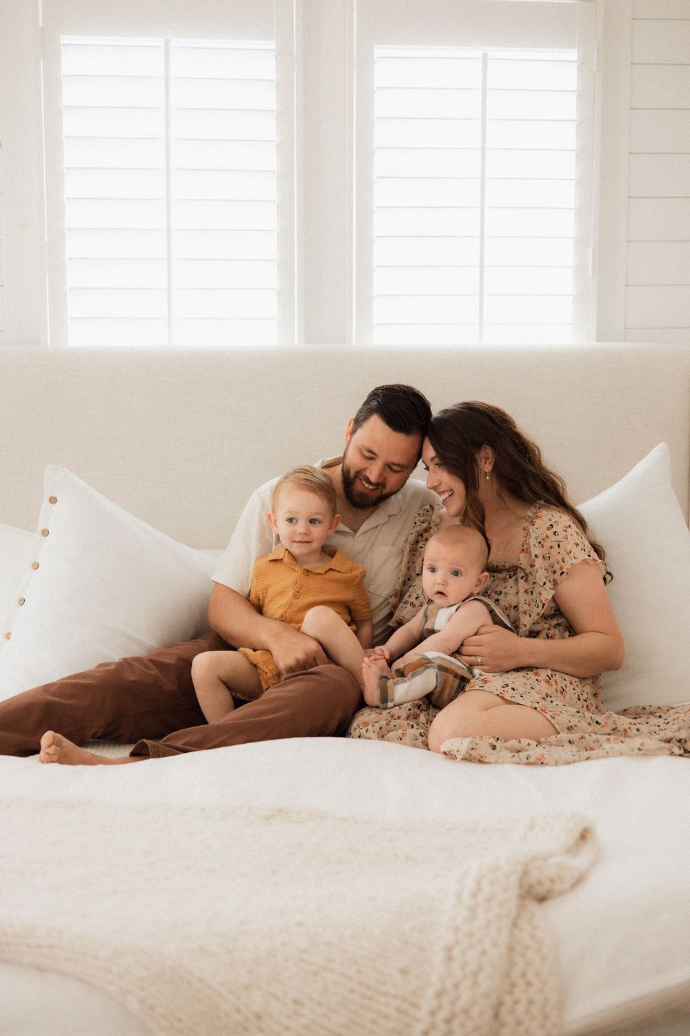 A family relaxes together on a white couch by bright windows, sharing a tender moment.
