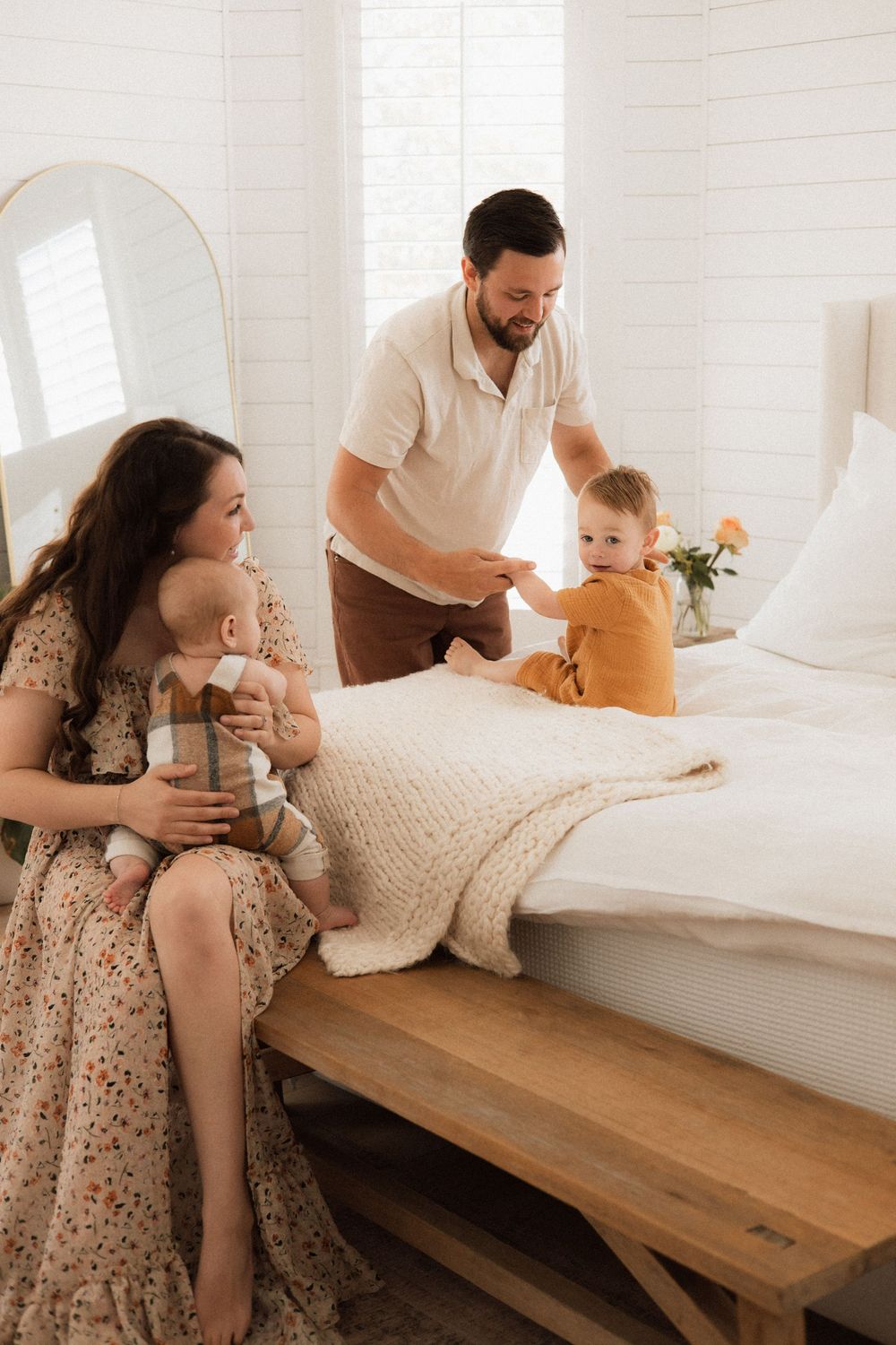 A family shares a tender moment together on a bed with white linens in a bright, minimalist bedroom setting.