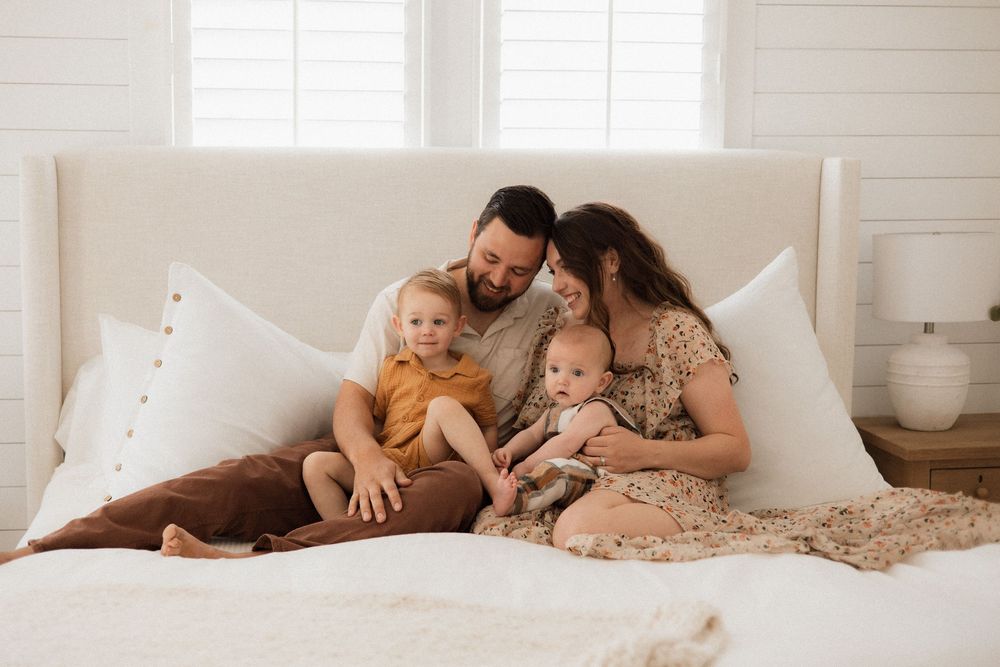 Family relaxes together on white bed with pillows and natural light streaming through window in bedroom.