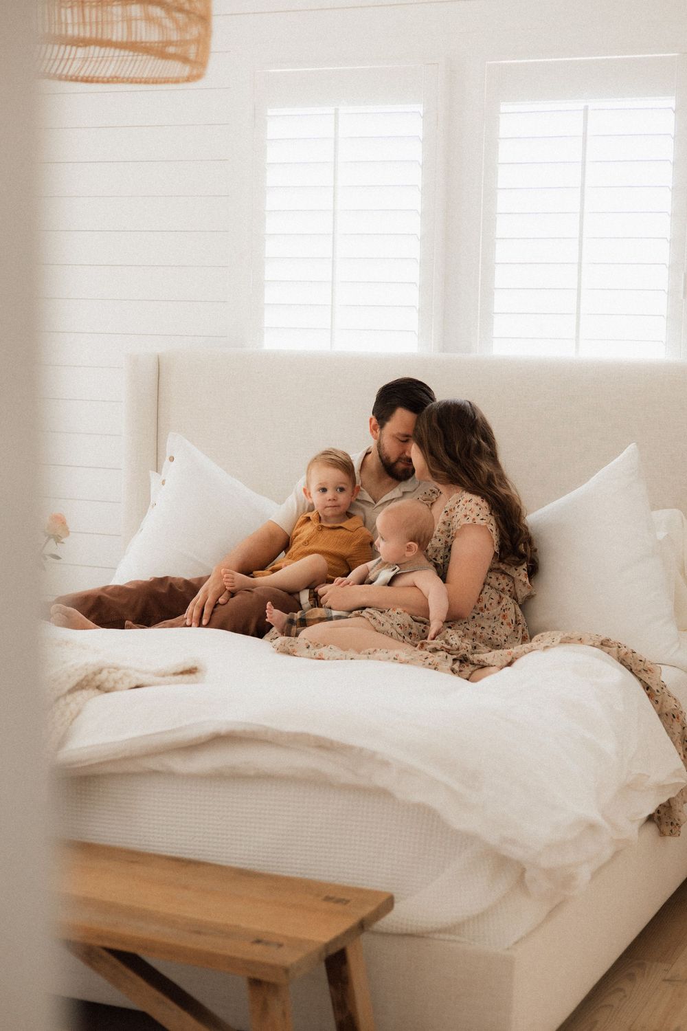 A family relaxes together on a white bed with natural lighting from windows and a woven pendant lamp above.