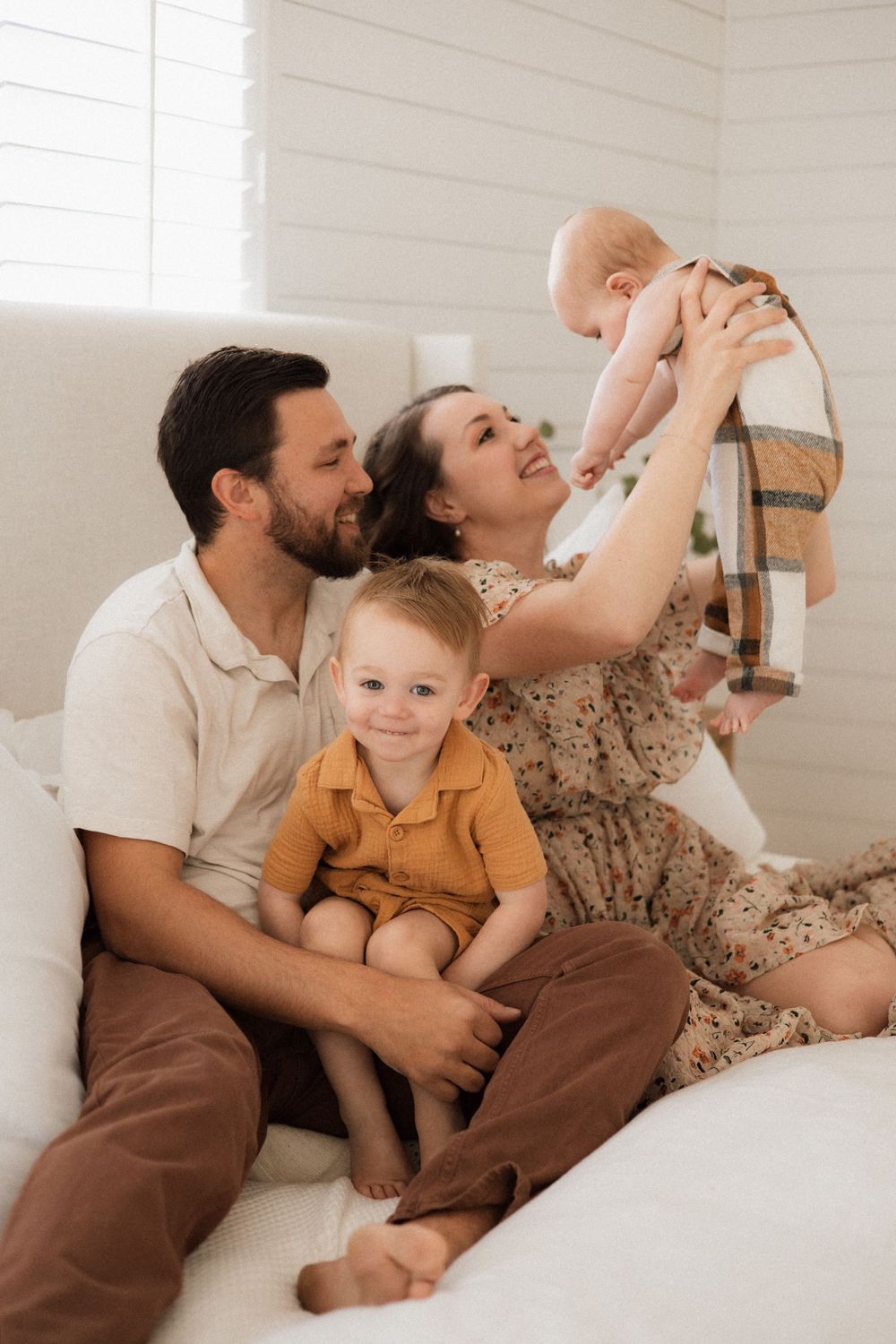 Family relaxes together on white couch while mother lifts baby in floral dress and father sits with toddler in mustard shirt.