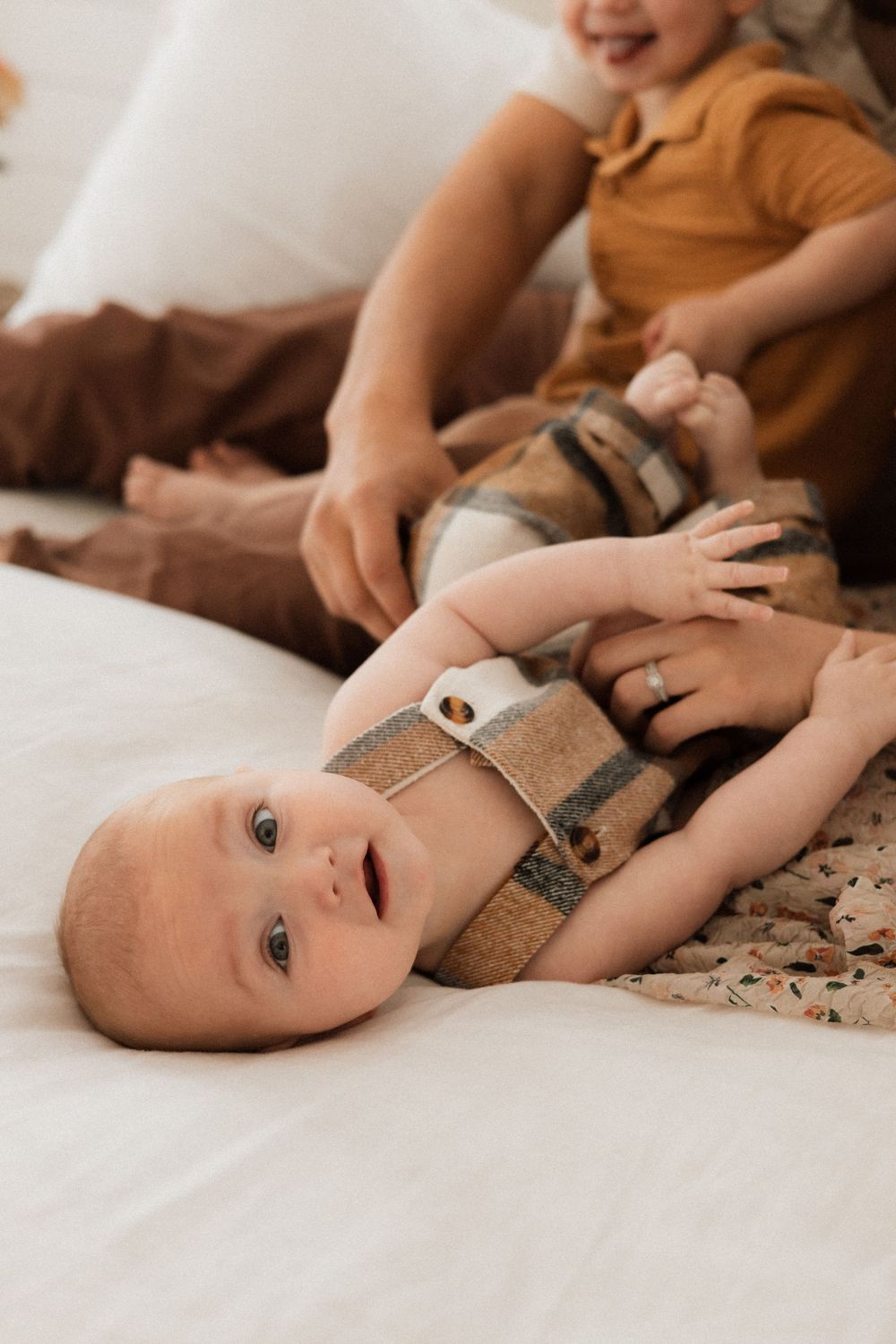 Baby wearing striped overalls lies on white bedding while being tickled.
