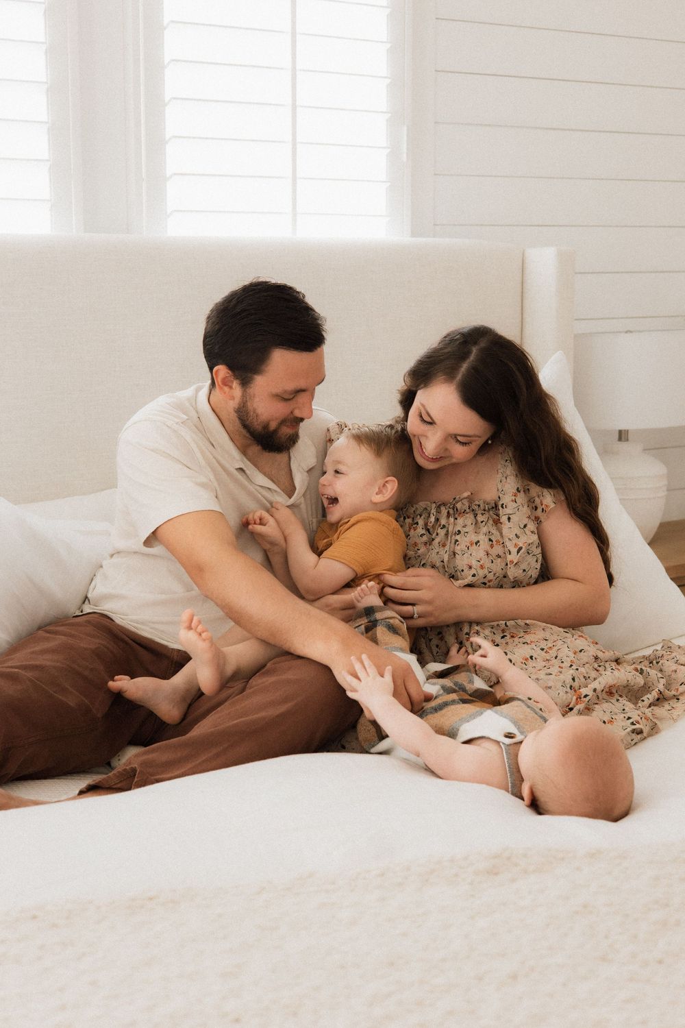 A tender family moment as parents and two young children share cuddles and laughter together on a white bed.