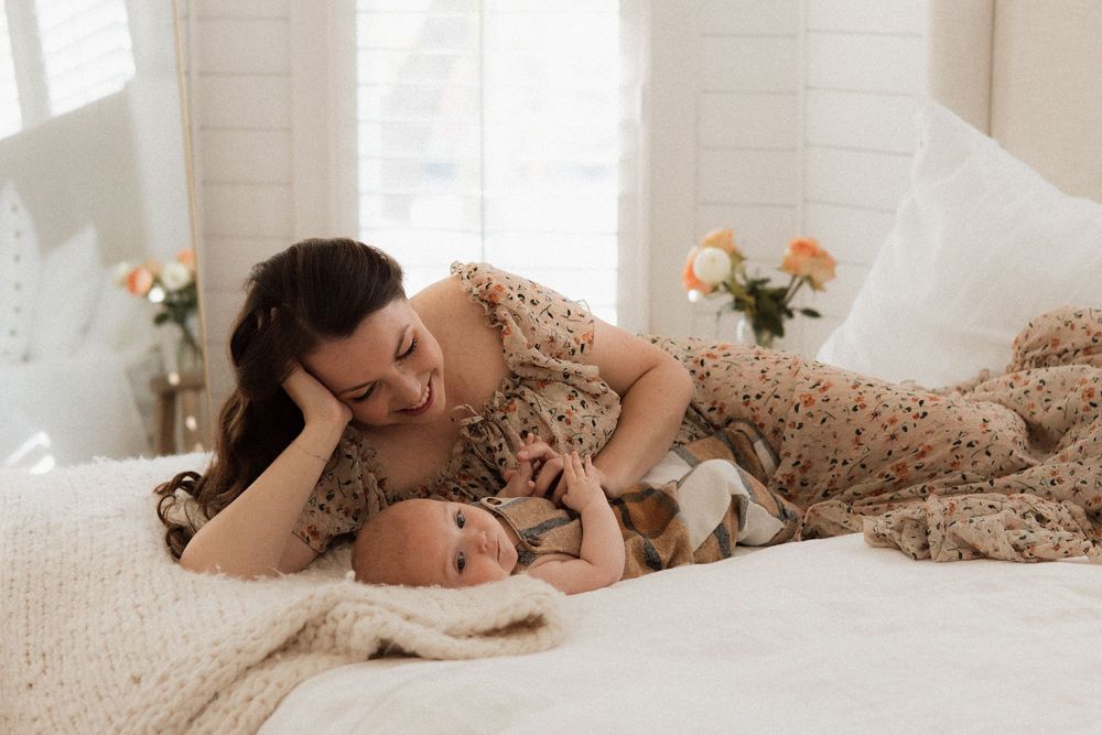 A parent lies on a white bed while tenderly caring for an infant under cozy beige blankets with flowers in background.