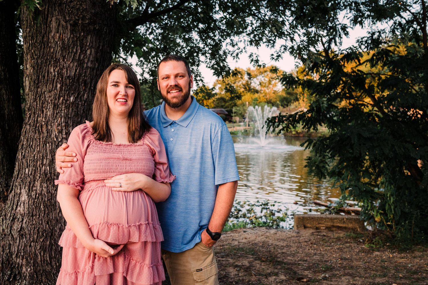 Expecting couple poses next to lake during sunset with trees in background and woman wearing pink maternity dress.