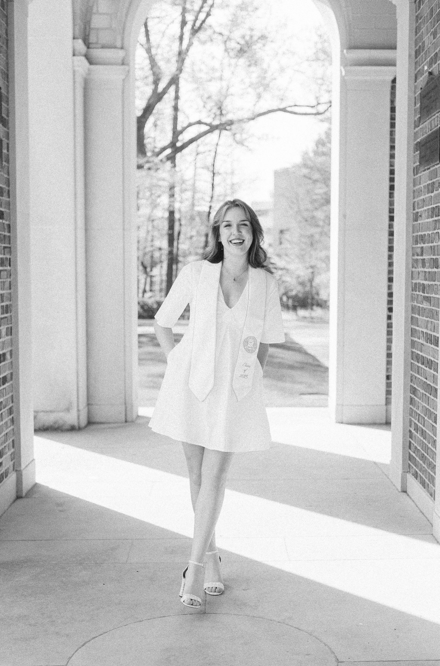 Candid portrait of a smiling college graduate wearing a white dress and stole, framed by the Bell Tower arches at UNC