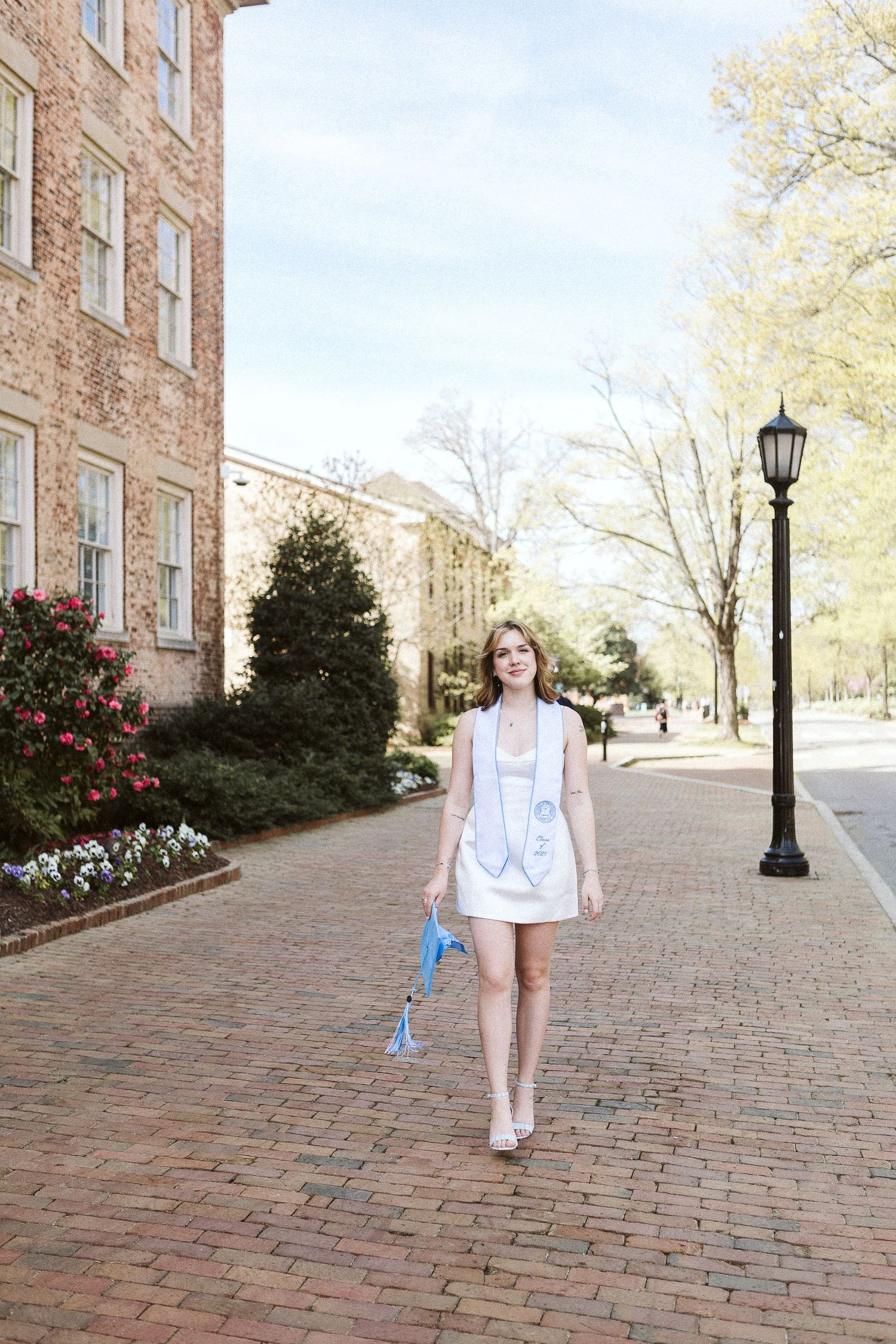 Natural light portrait of UNC graduate walking with a soft smile along the sidewalk near the Old Well