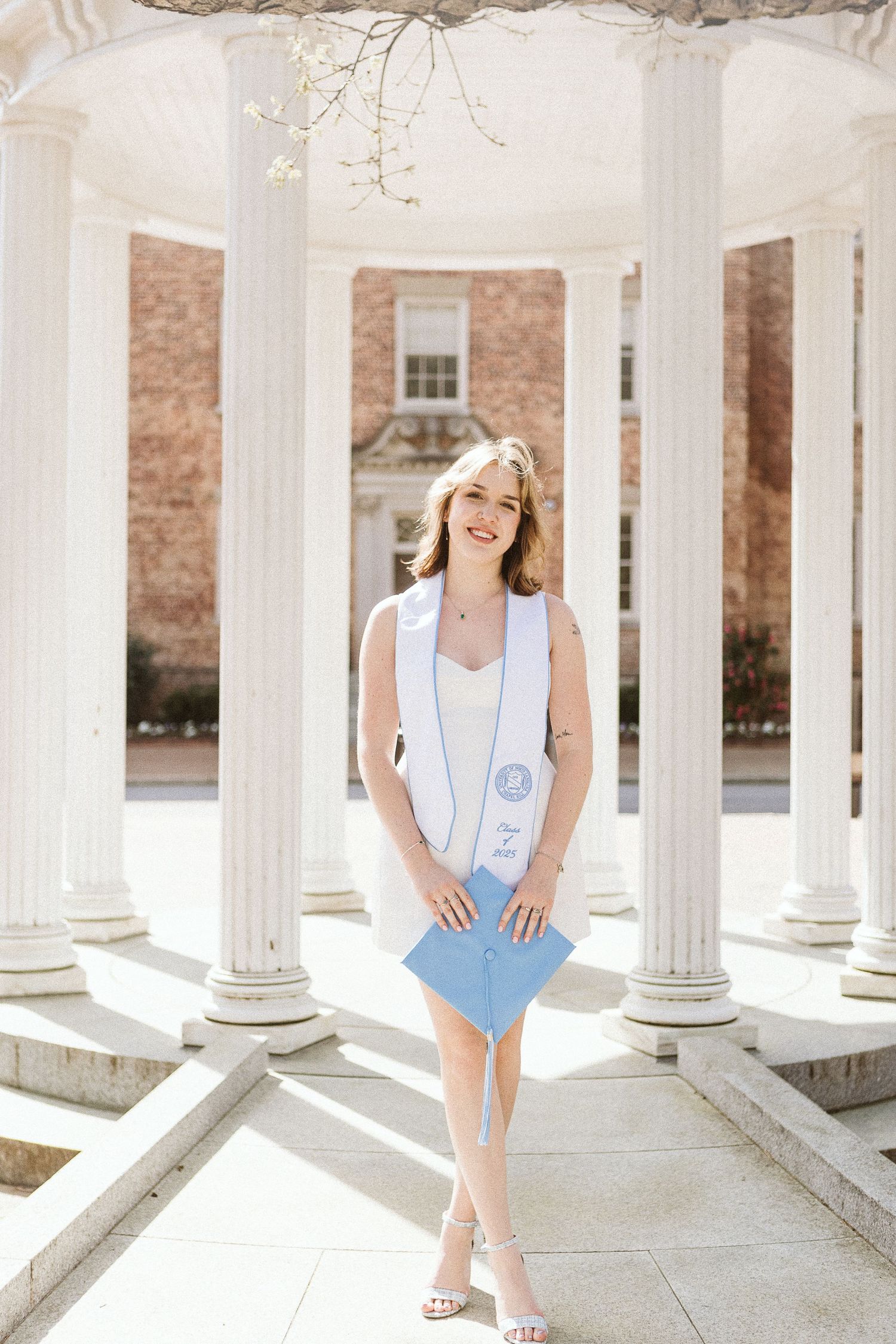 UNC Chapel Hill graduate in white dress and stole standing in front of the iconic Old Well in natural light