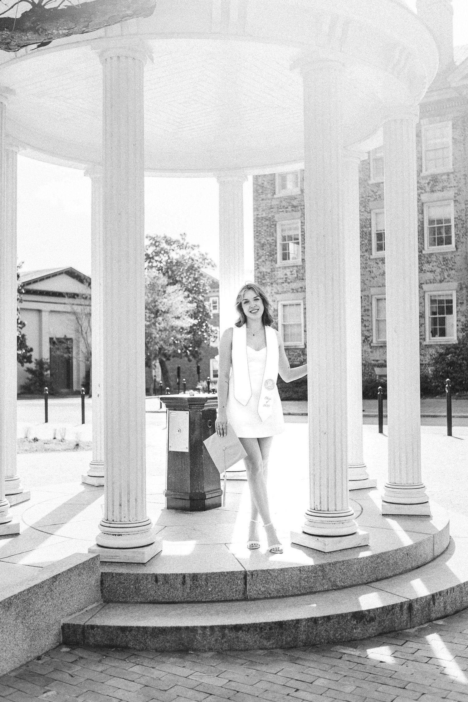 Artistic black and white portrait of a graduate in white dress and stole standing in front of the iconic Old Well in natural light