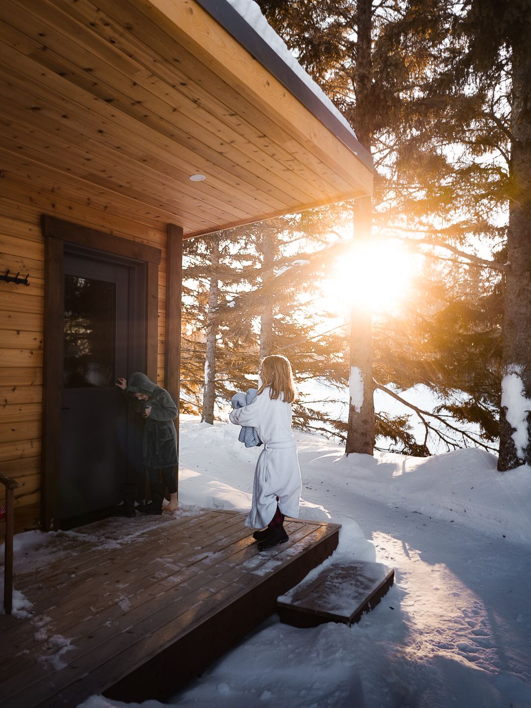 Sunlit wooden cabin deck covered in snow with golden hour light streaming through pine trees.