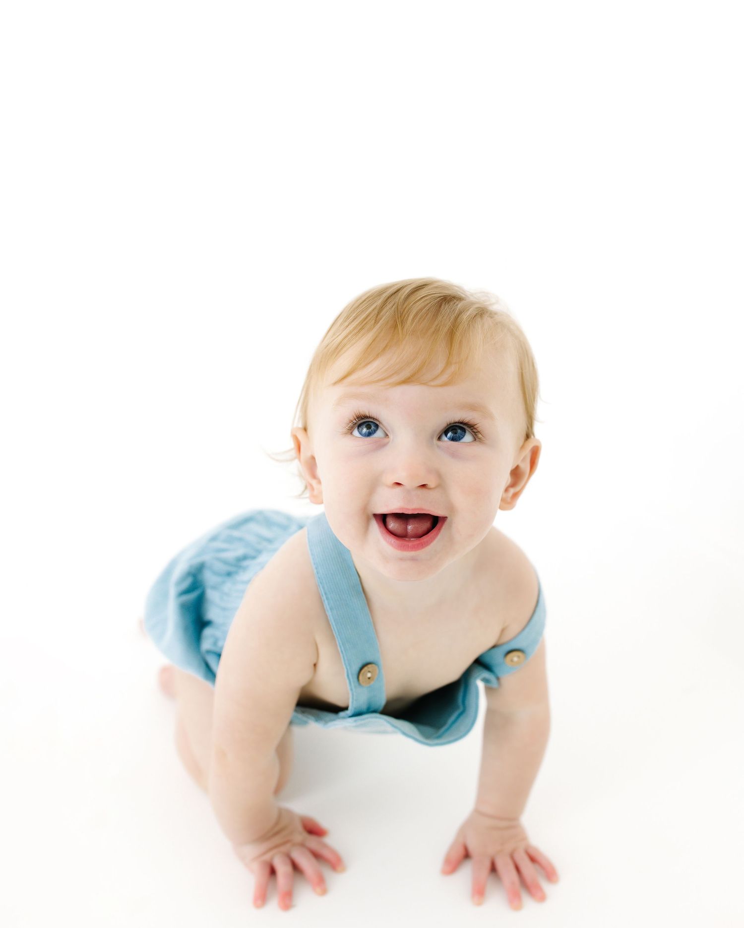 A joyful baby in a light blue outfit crawls on a white surface while looking up with bright eyes and a big smile.