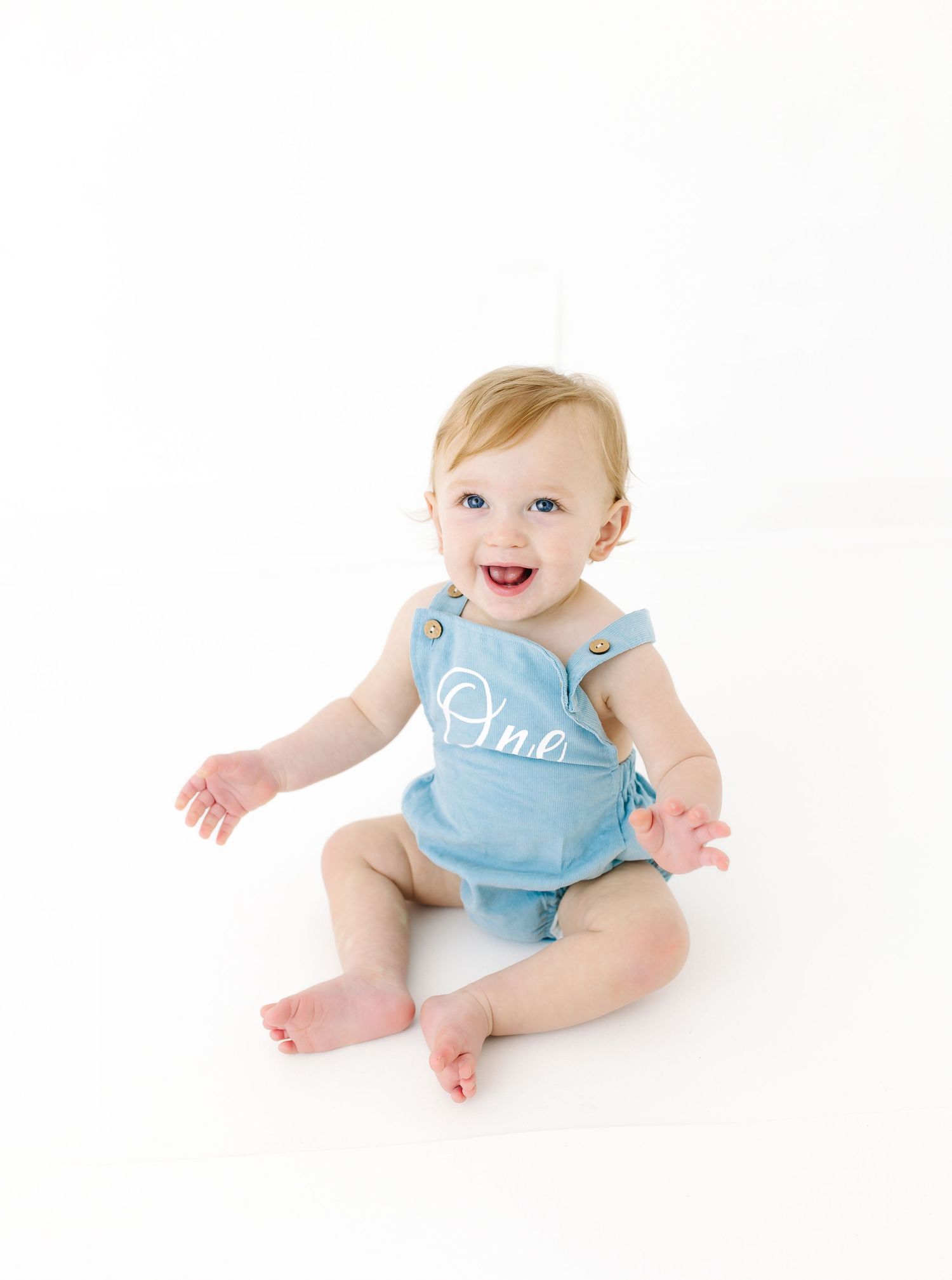 Baby wearing light blue romper with white lettering sits on white background with joyful expression.