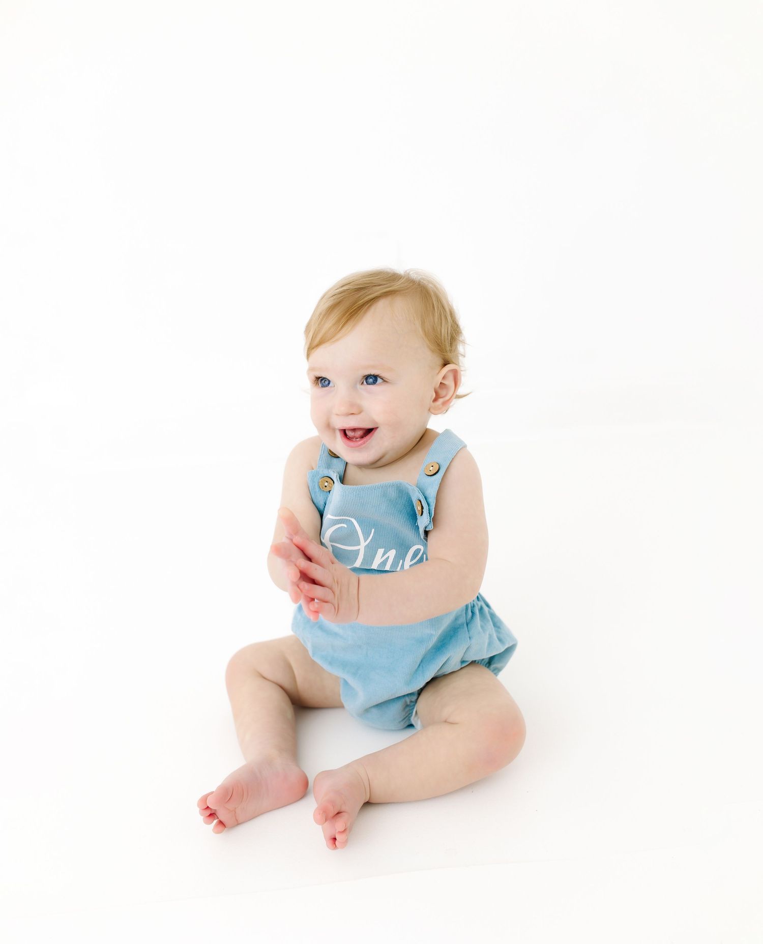 Baby in blue sleeveless romper laughs while sitting against white background.