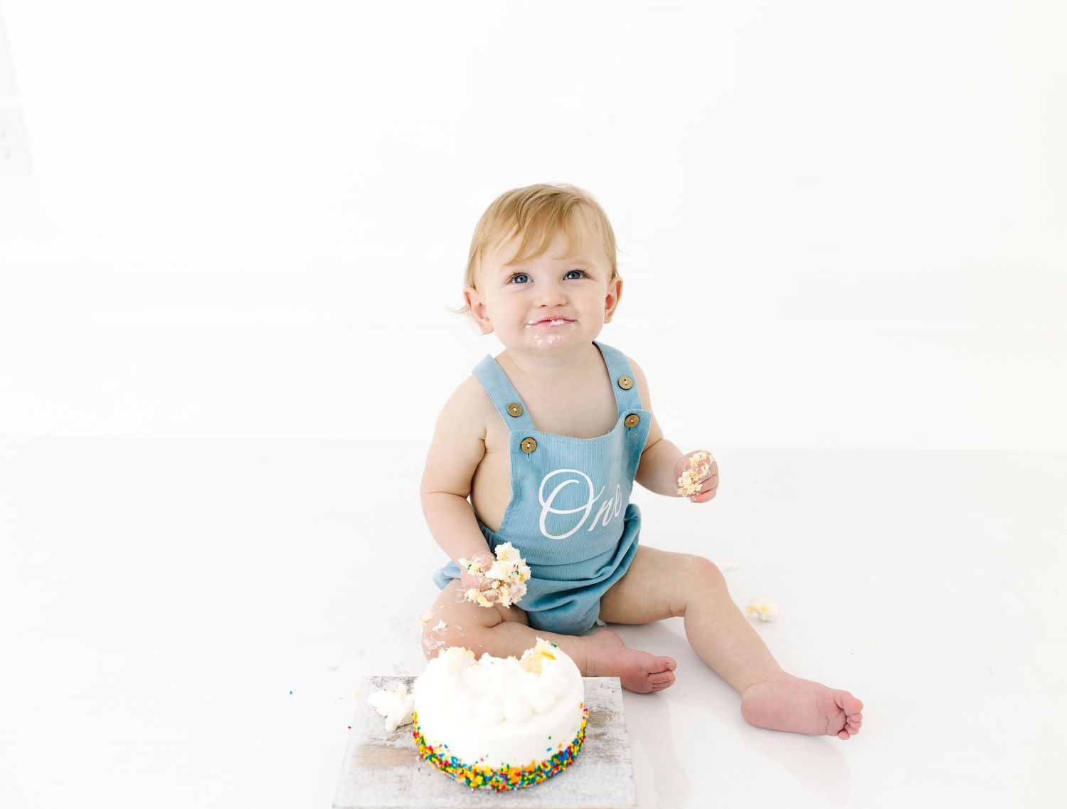 Baby wearing blue romper sits on white background with frosted cake during first birthday photo session.