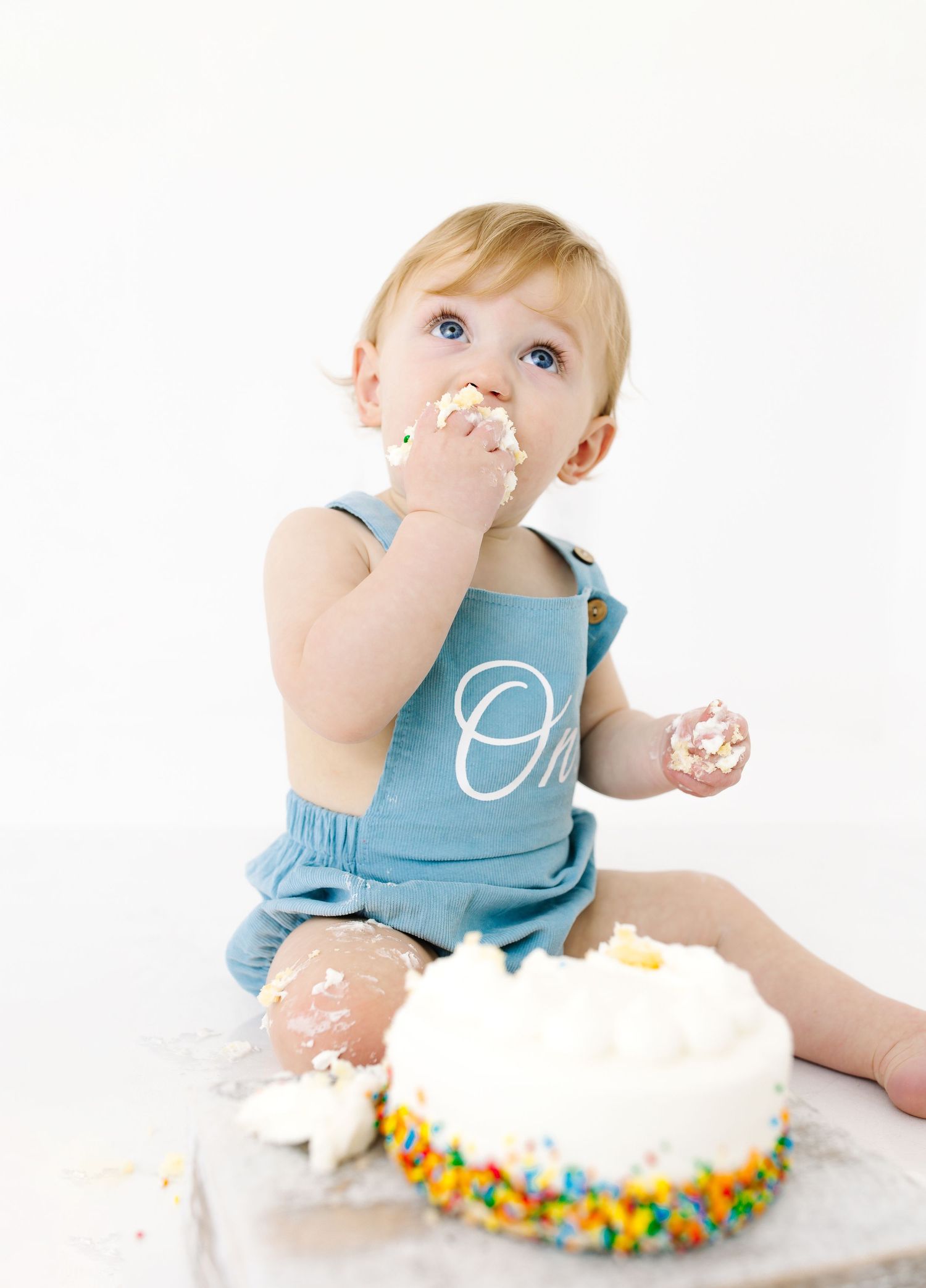 Small child in blue overalls enjoys a white birthday cake with colorful sprinkles against a white background.