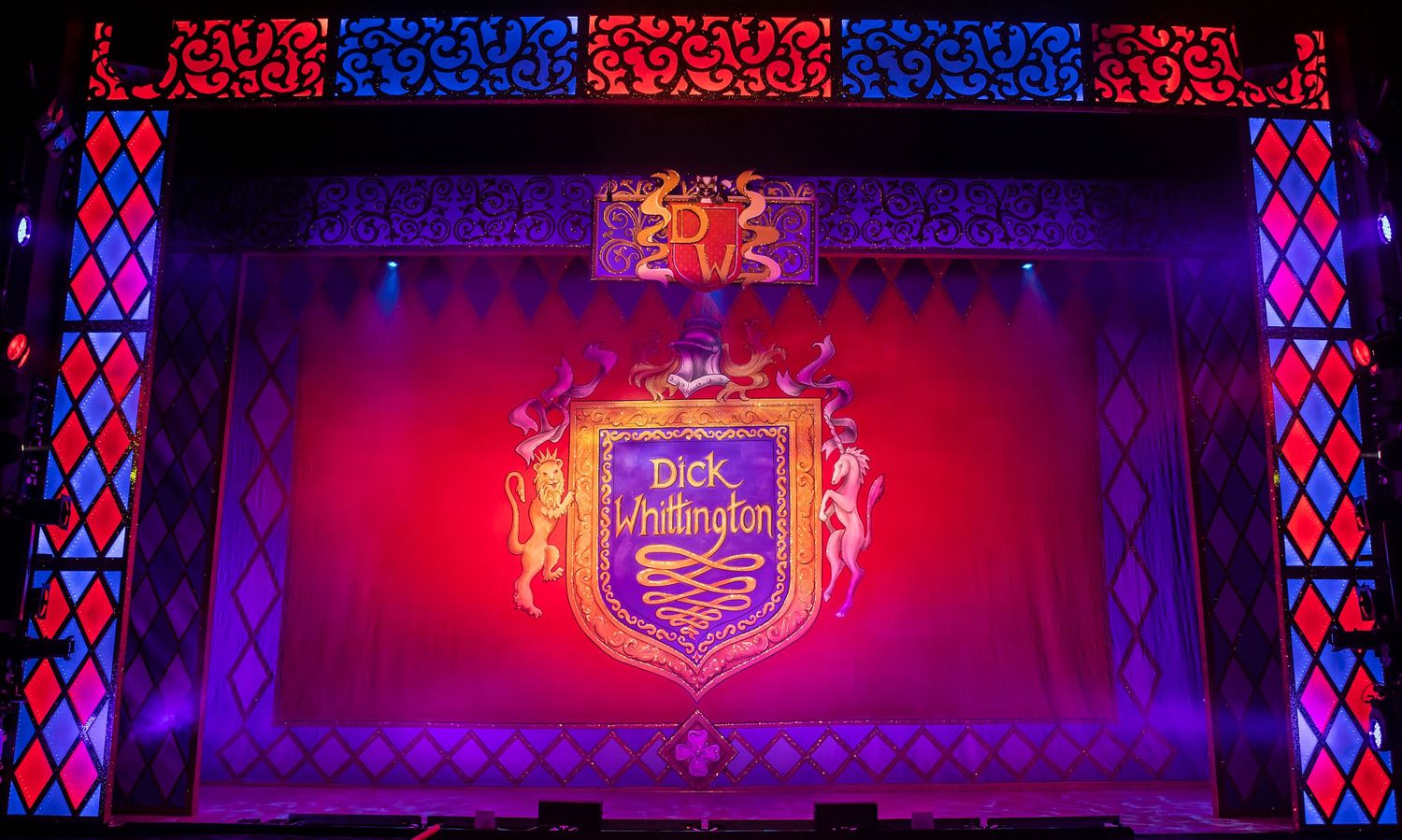 Theater backdrop displaying a stylized medieval crest design illuminated in red and blue theatrical lighting.