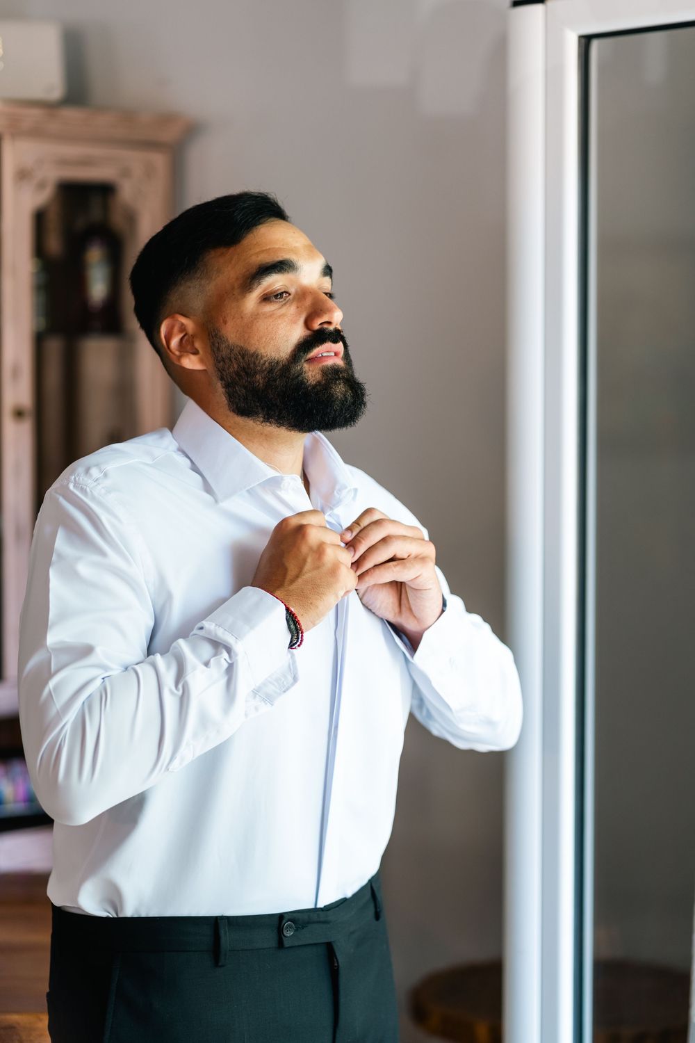 A person adjusting a white dress shirt while getting ready near a window.