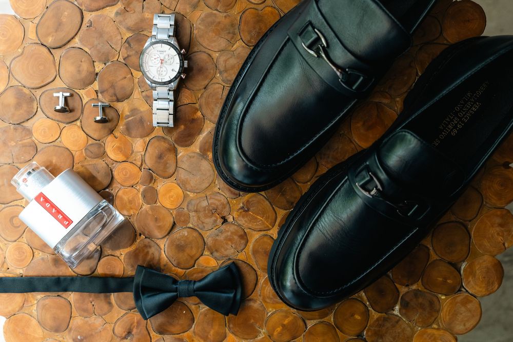 Wedding accessories including black leather shoes, watch, and bow tie arranged on wooden surface.