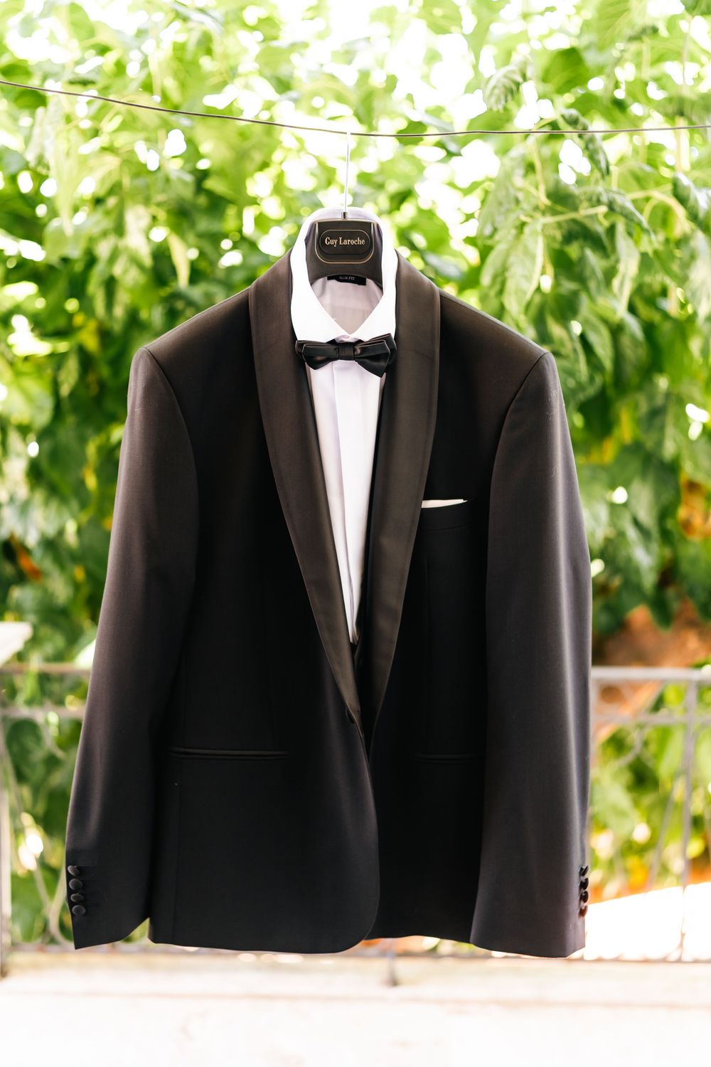 Classic black tuxedo jacket with bow tie hangs on display against lush green foliage.