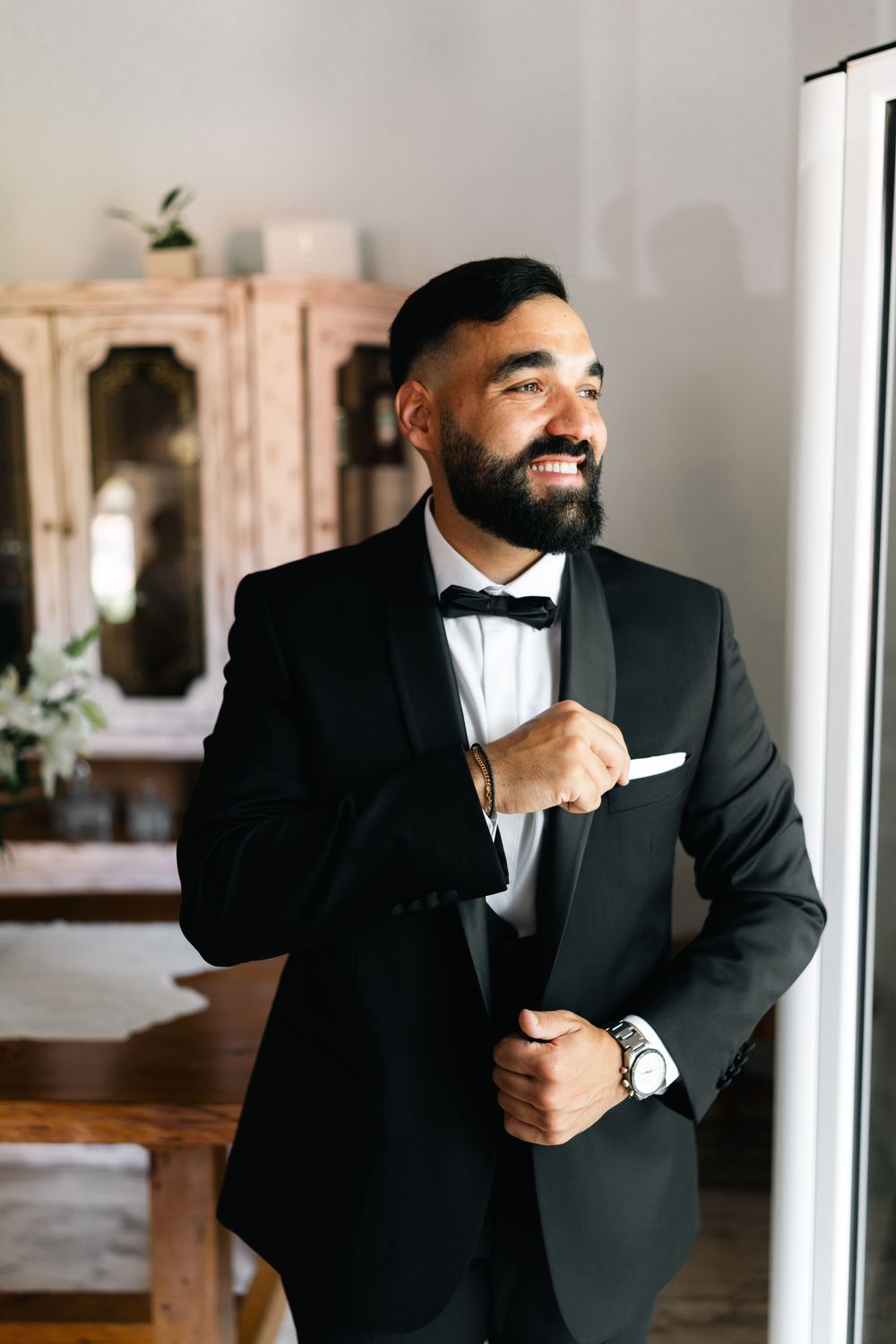 Person in formal black tuxedo and bow tie standing in elegant interior setting.