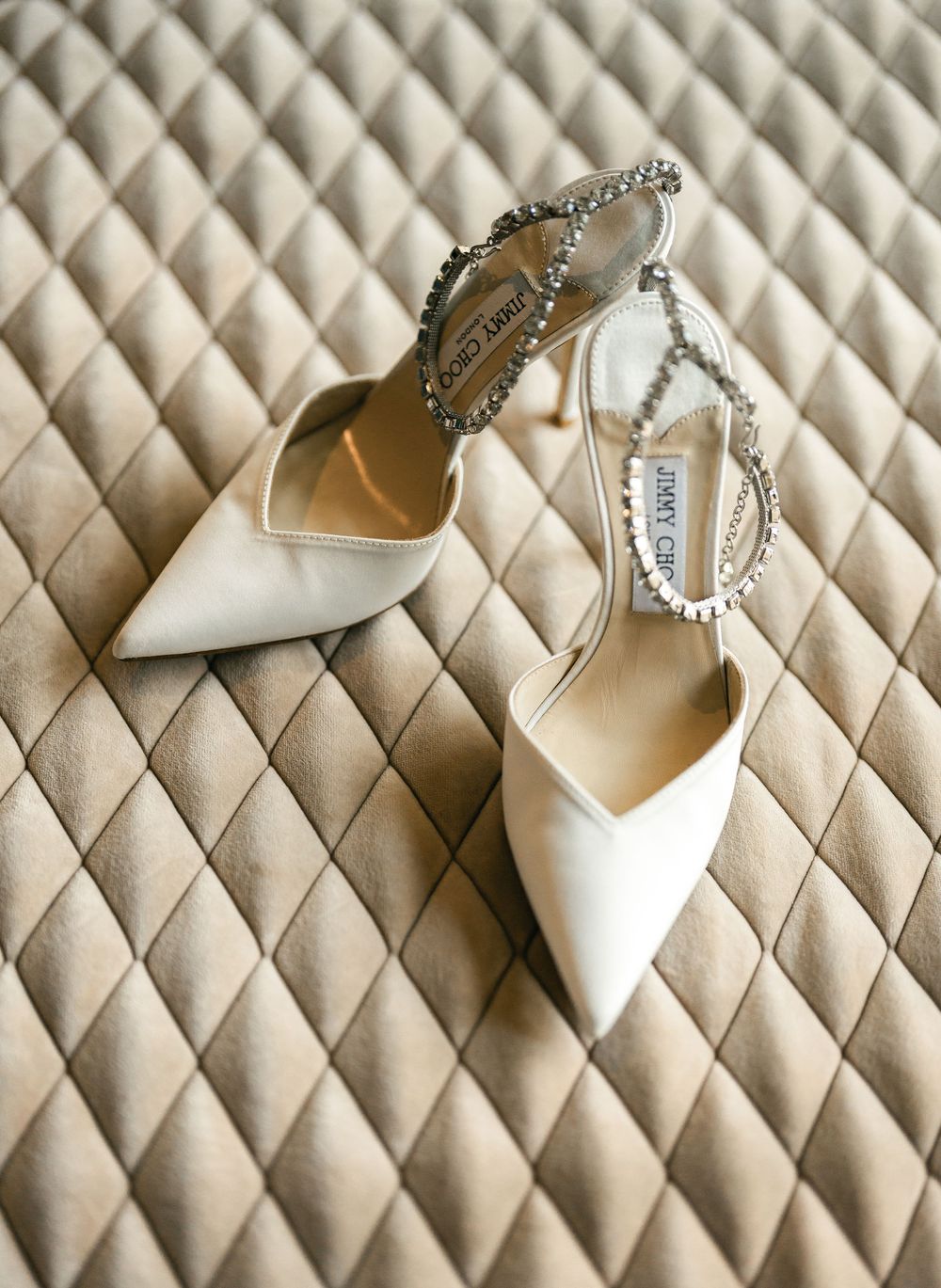 Elegant white bridal heels with crystal embellished ankle straps lay on quilted cream fabric.