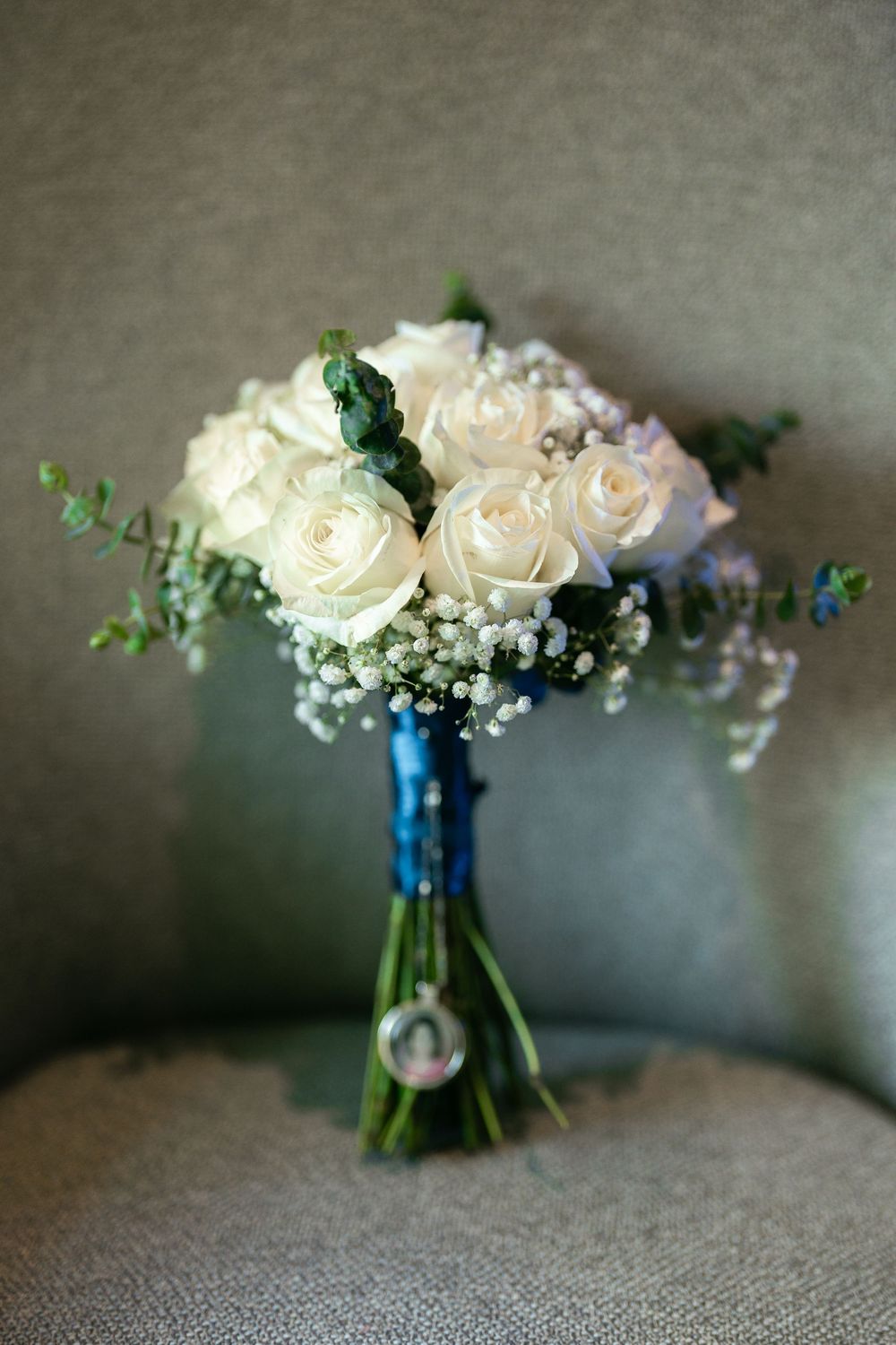 Elegant bridal bouquet with cream roses, baby's breath and eucalyptus tied with blue ribbon on gray textured background.