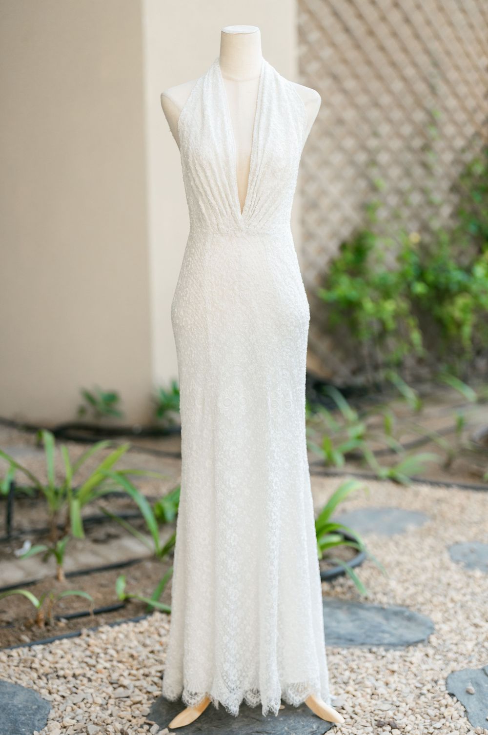 Elegant white lace wedding gown with plunging v-neck and fitted silhouette displayed on mannequin in garden setting.
