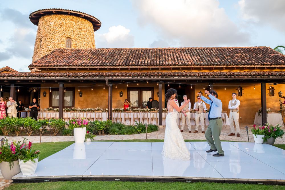 Wedding couple dancing on white platform in front of rustic Spanish-style building with tower.