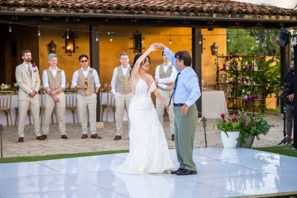 Bride and groom share first dance with groomsmen watching on outdoor dance floor.