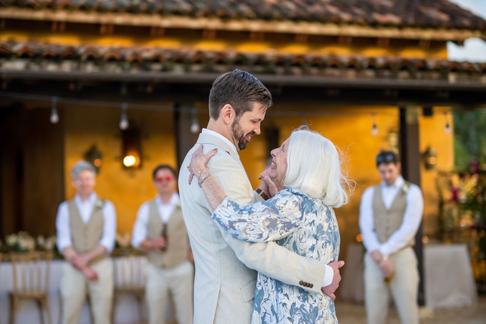 Intimate dance moment at a wedding reception with Spanish-style yellow building and groomsmen in background.
