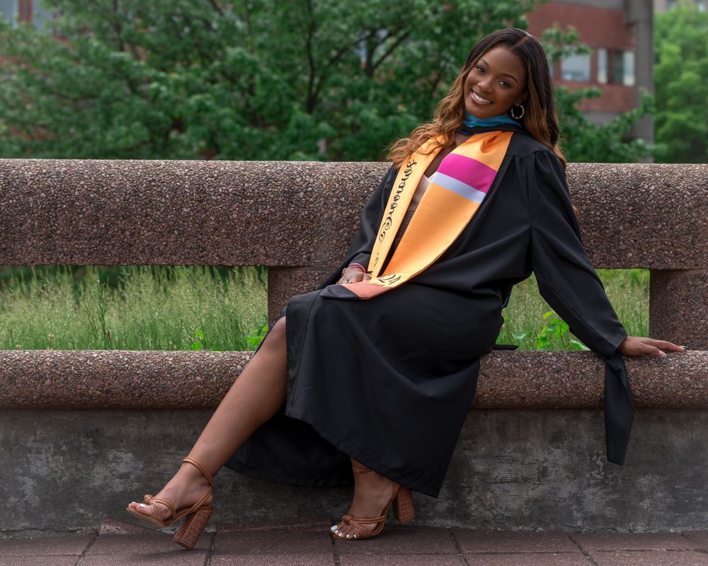 Graduate in academic regalia relaxing on stone bench during outdoor photo session.