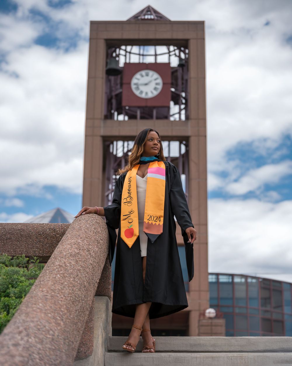 Graduate in cap and gown posing in front of clock tower wearing decorative graduation stole.