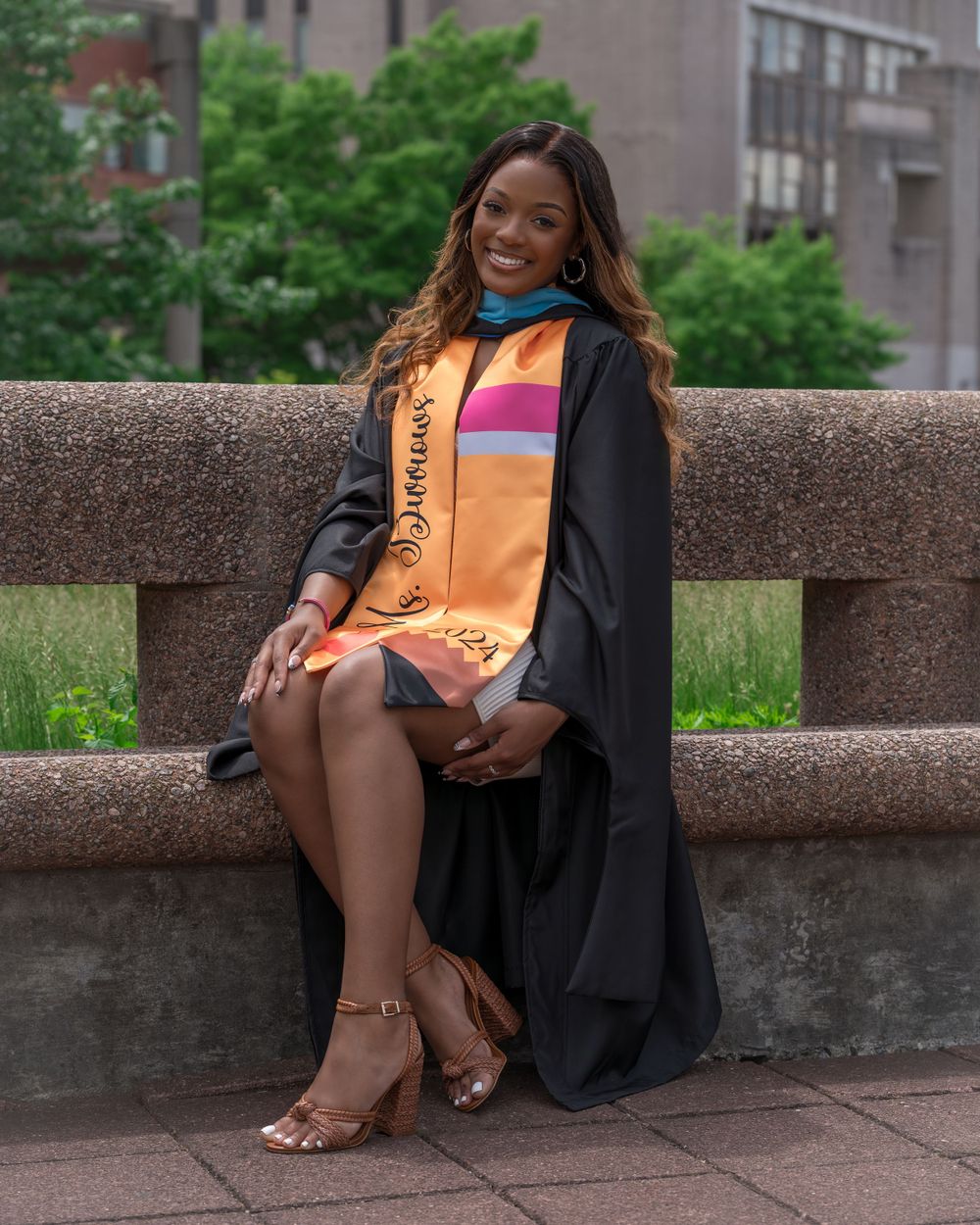 Graduate in academic regalia sitting on stone wall with colorful graduation stole.