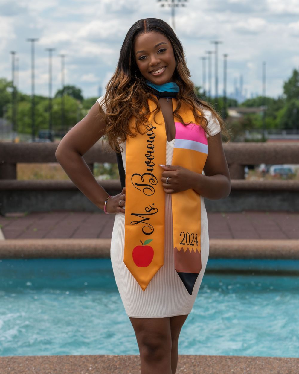 Graduate in white dress and orange sash poses by fountain in celebratory portrait.