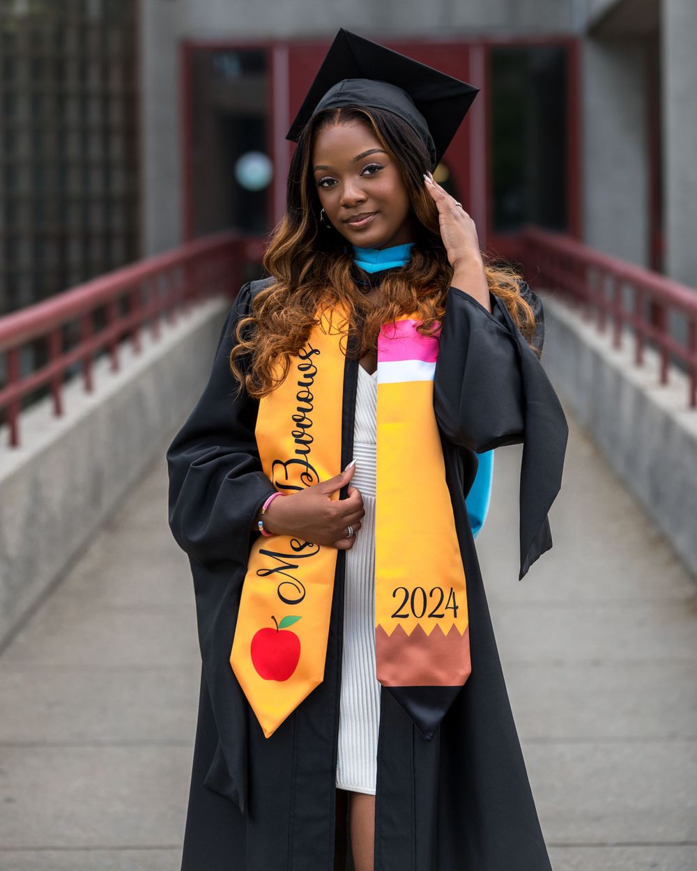 Graduate in cap and gown with decorative 2021 sash poses on bridge.