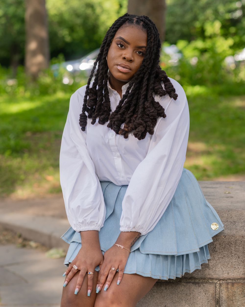 A person in a white blouse and light blue pleated skirt sits on stone steps outdoors.
