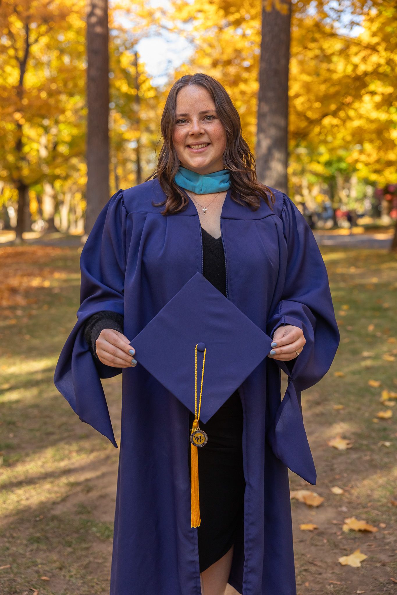 Cap and Gown Session Graduate in navy blue academic regalia holds diploma in autumn setting with golden leaves. Spokane senior photographer.