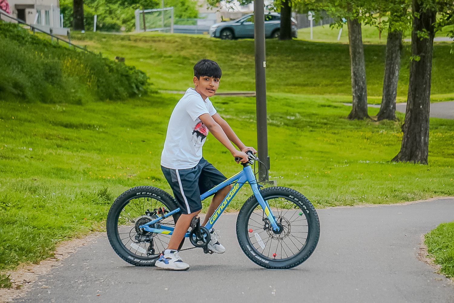 A person rides a turquoise fat tire mountain bike on a paved path through a green park setting.