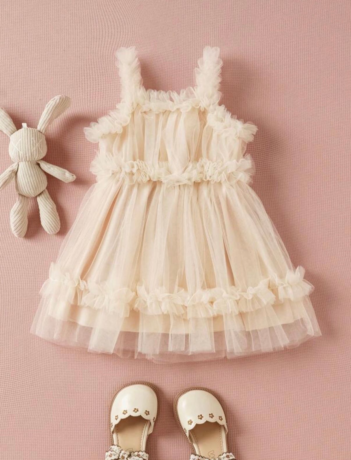 Delicate cream-colored lace dress with ruffled details and matching accessories laid on pink background.